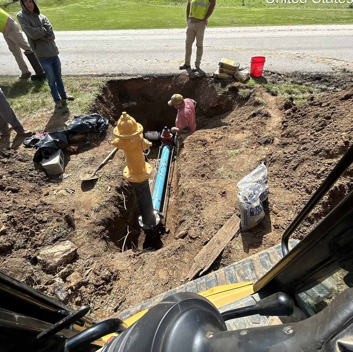 Workers repairing a water line by a road, with a fire hydrant, blue pipe, and red bucket.