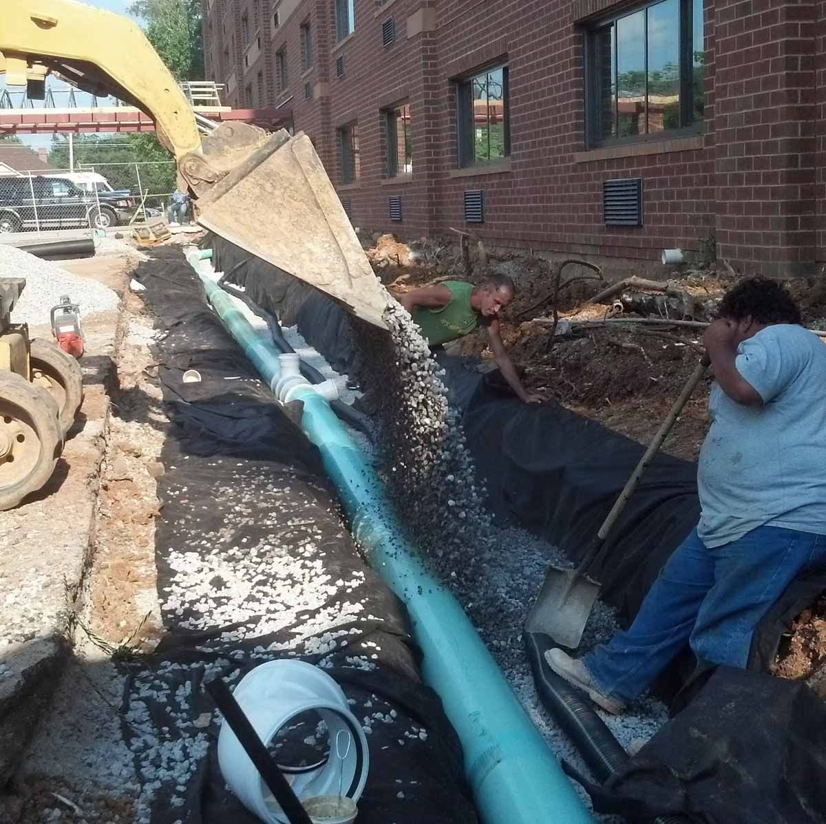 Construction workers backfilling a trench around a blue pipe near a brick building.