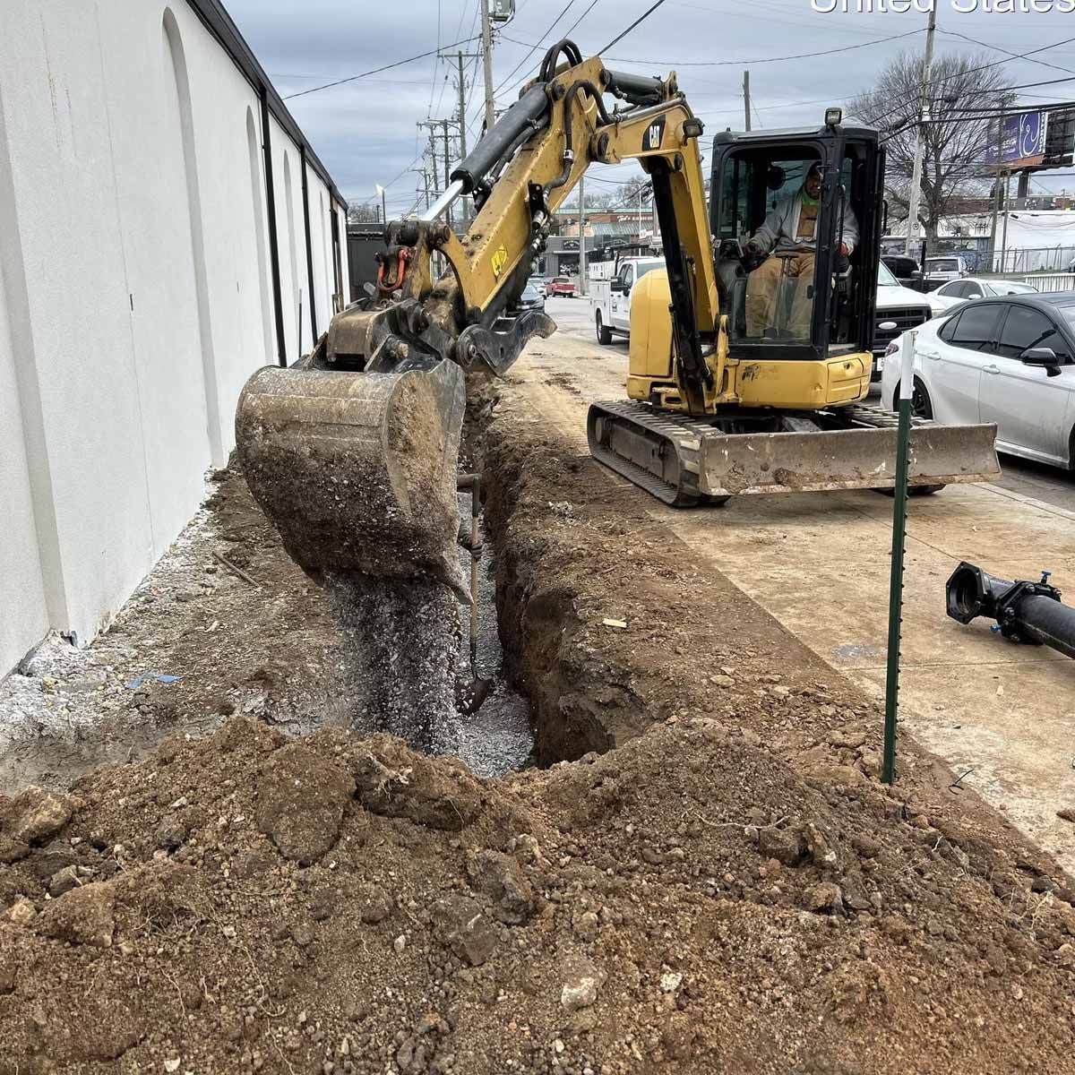 A yellow excavator digs a trench next to a building on a street.