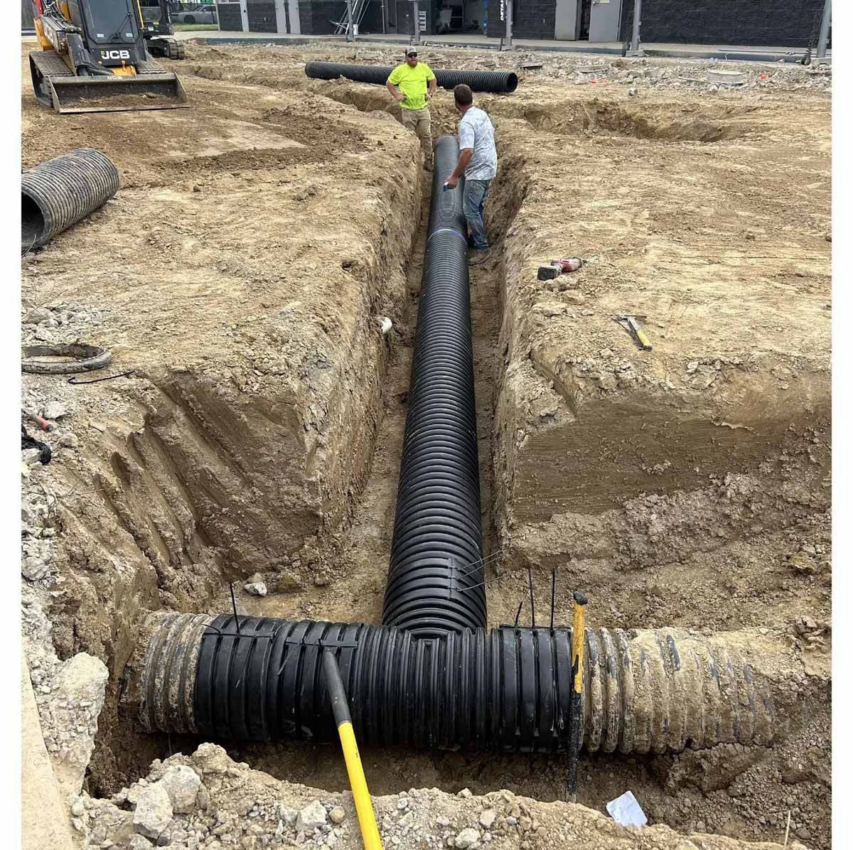 Workers install a black corrugated drain pipe in a dirt trench.