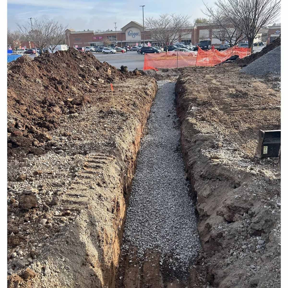 Trench filled with gravel, presumably for drainage, in front of a store and parking lot.