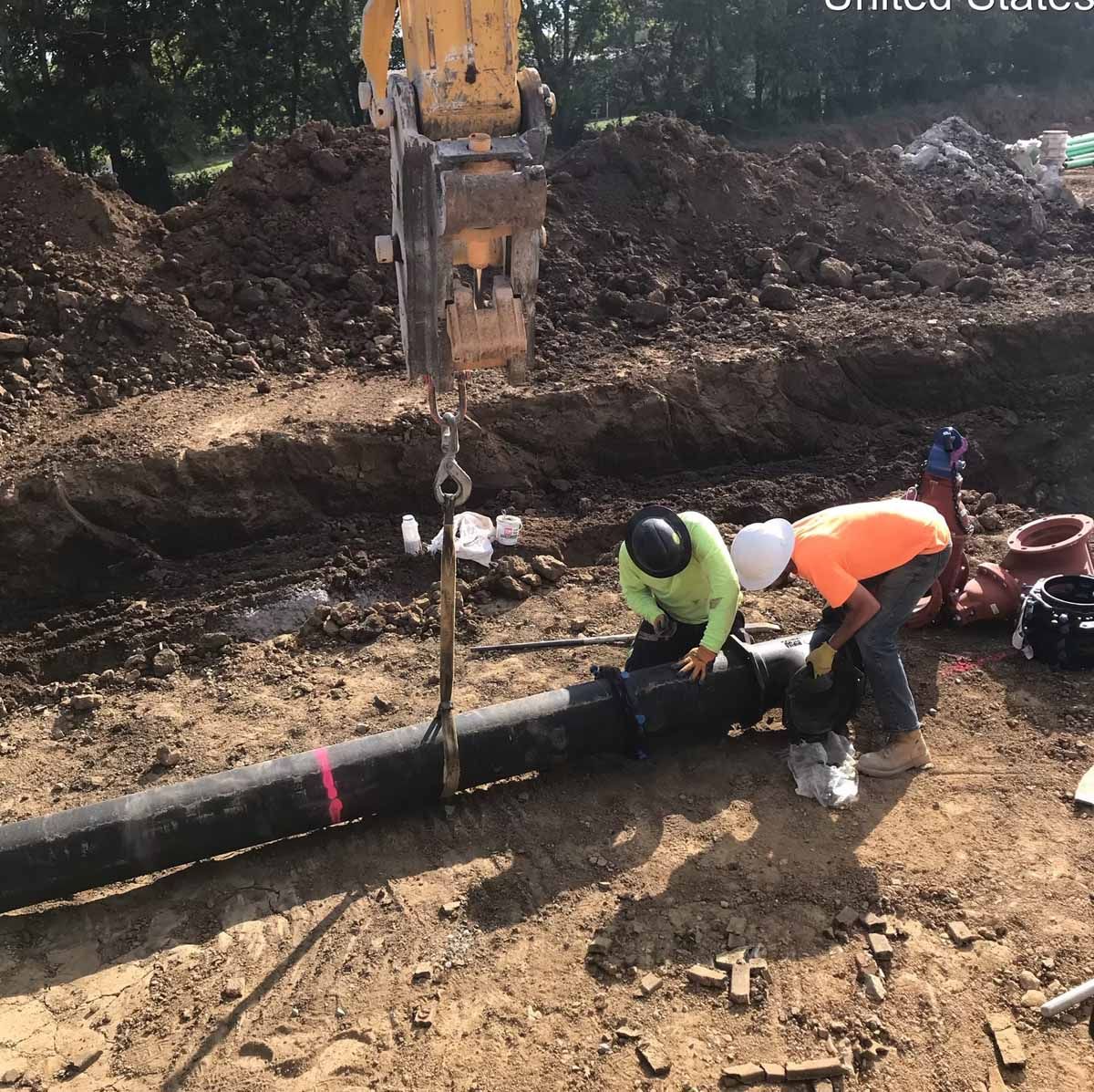 Construction workers connecting a large black pipe in a dirt trench, assisted by heavy machinery.