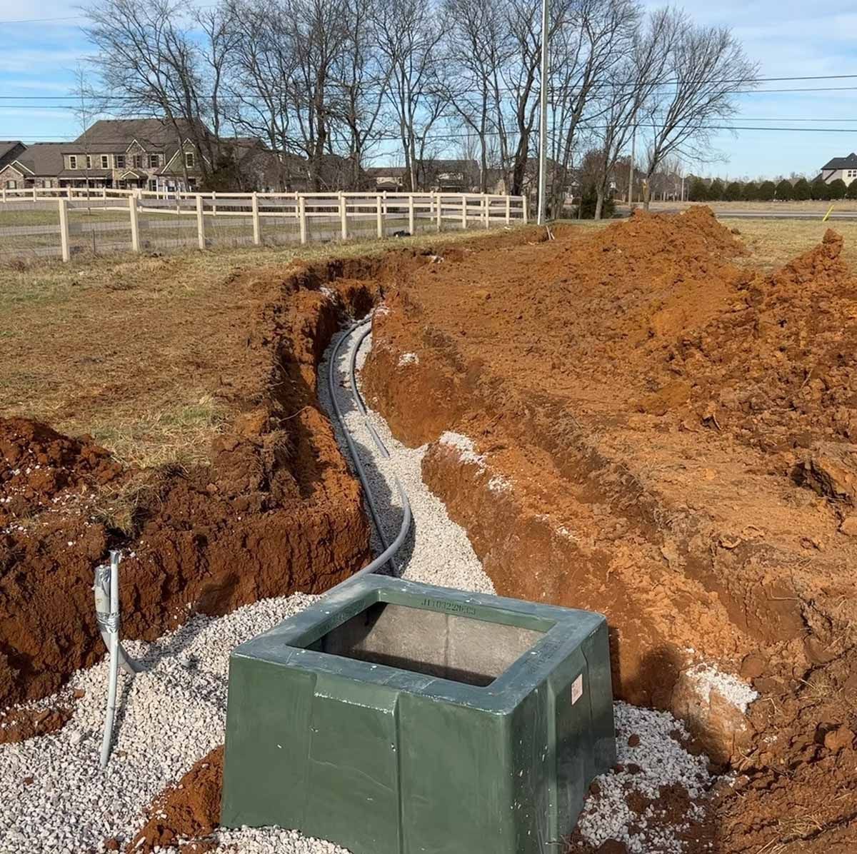 Trench with gravel and pipe, leading to a green square box. Dirt pile and field in the background.