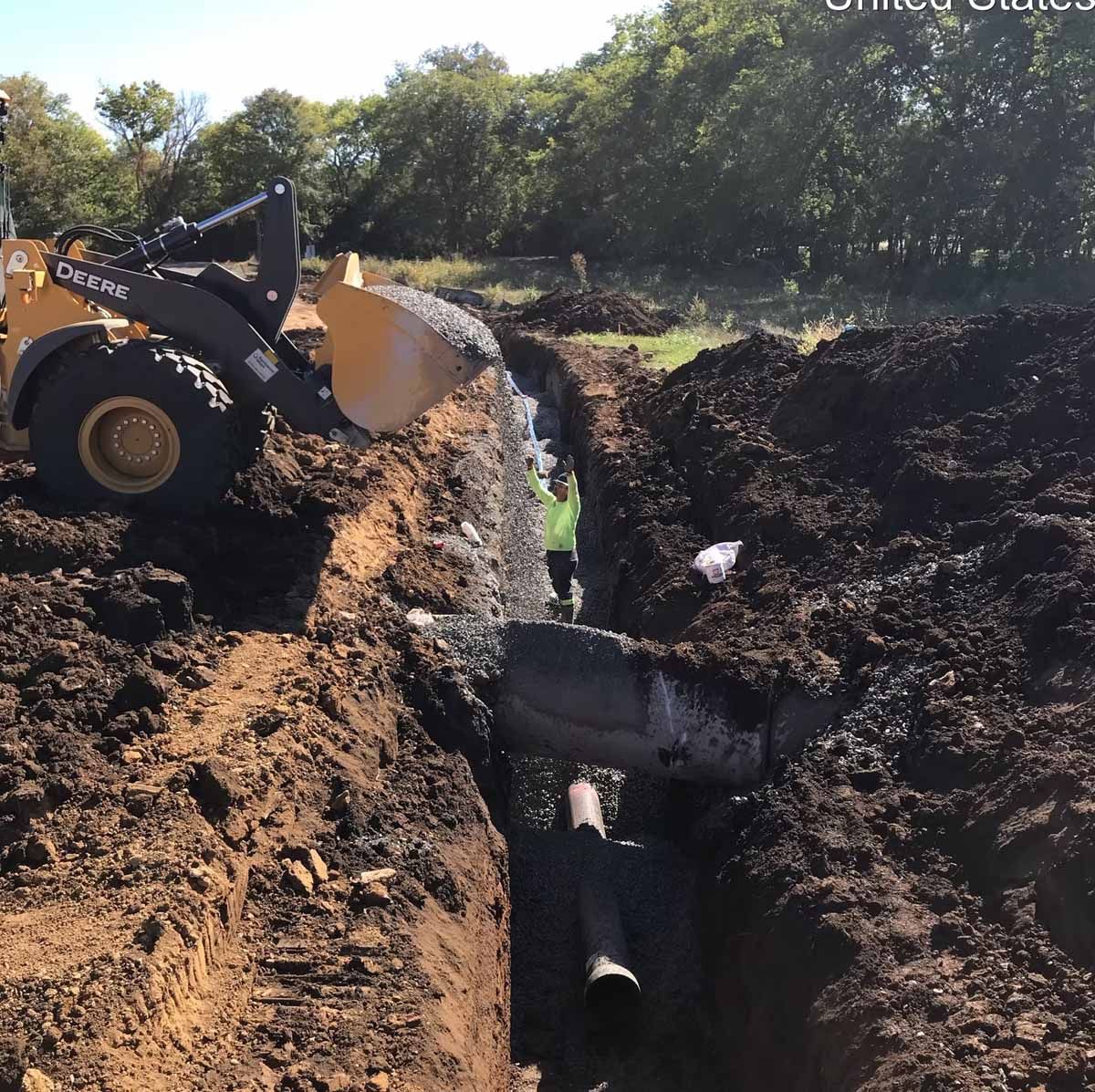 A yellow construction vehicle scoops dirt from a trench where a worker is inspecting a large pipe.