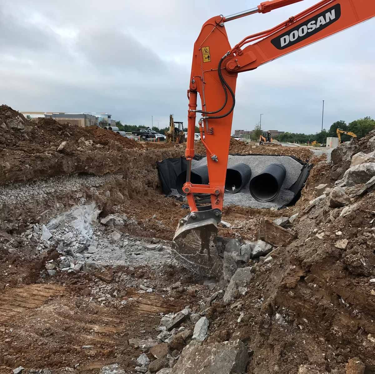 Orange excavator breaking up concrete in an excavation pit, with black pipes visible.