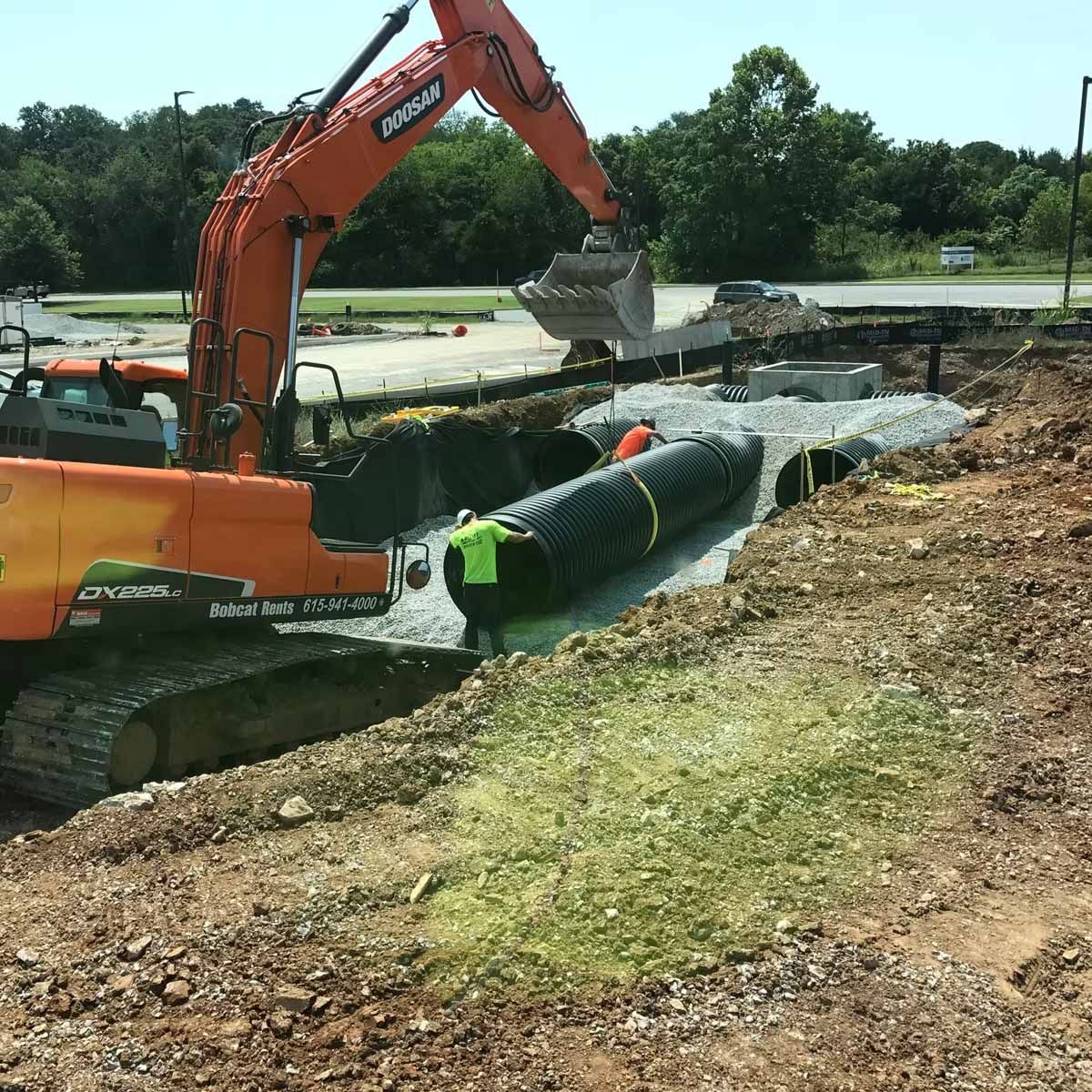 An orange excavator placing a black drainage pipe into a trench at a construction site.