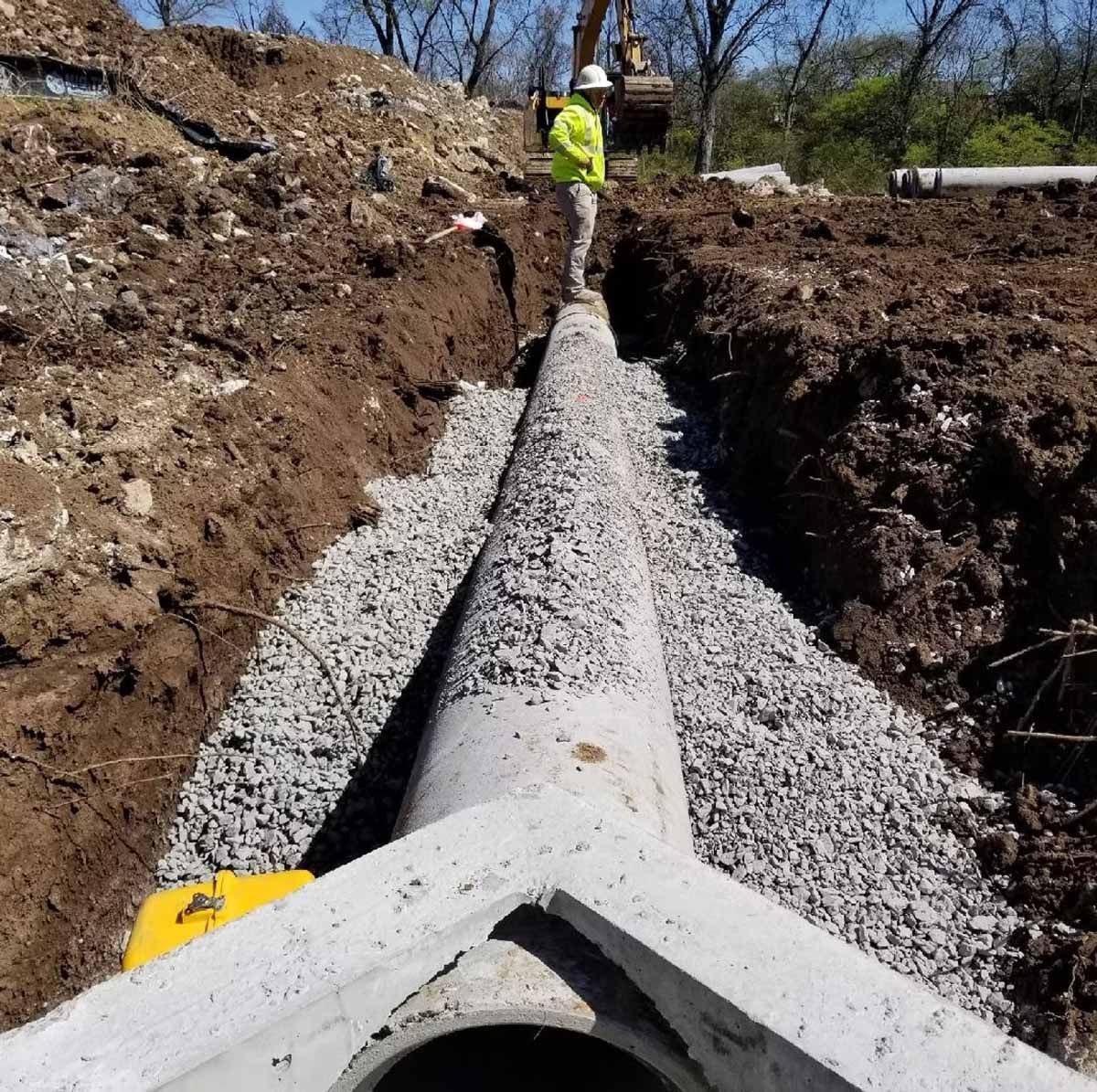 Construction of a concrete drainage pipe in an open trench, worker in yellow vest, outdoor setting.