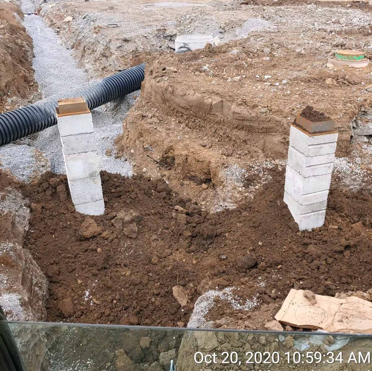 Construction site: two concrete block pillars in excavated dirt, near a drainpipe and gravel trench.
