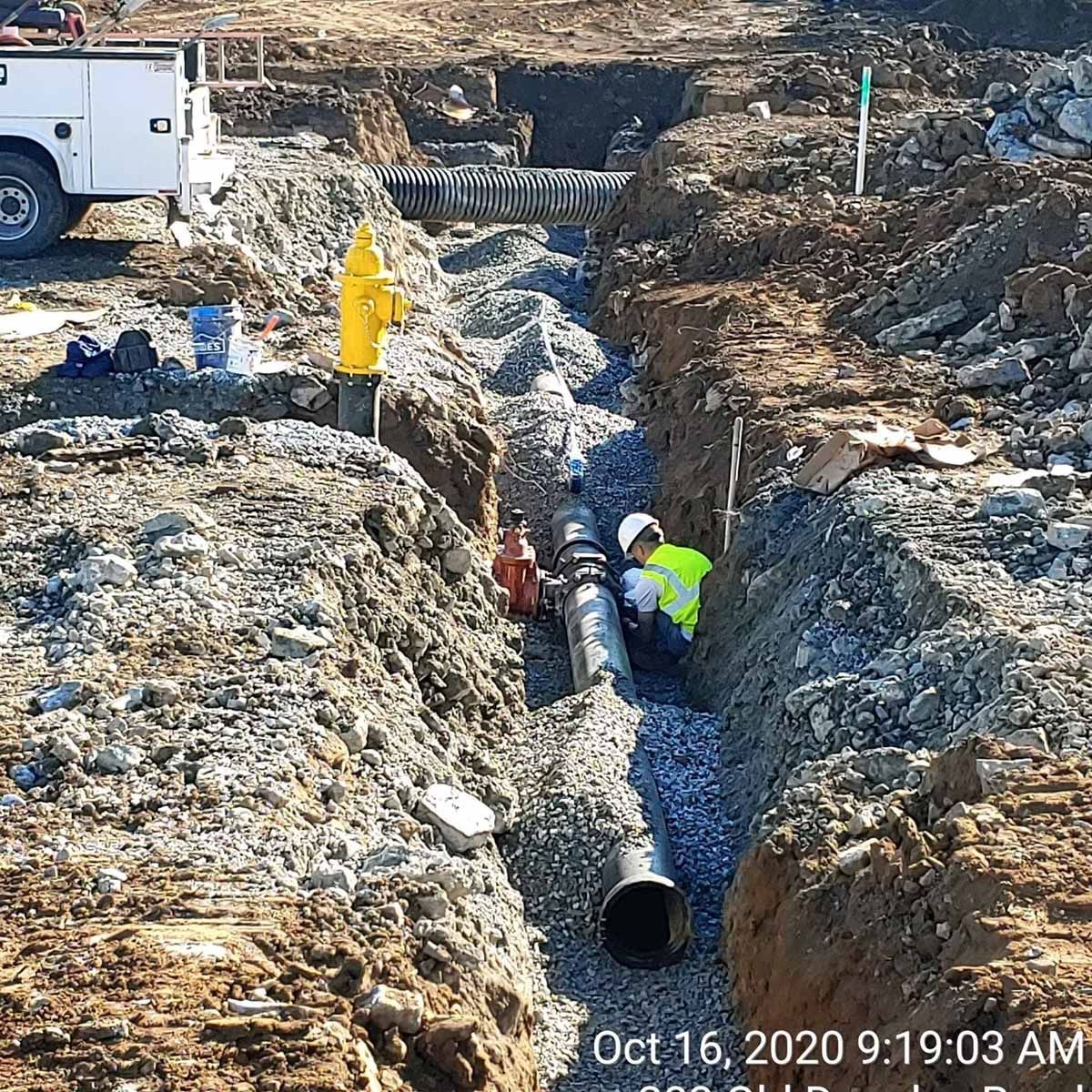 Construction workers installing a pipe in a trench. A yellow fire hydrant and truck are visible.