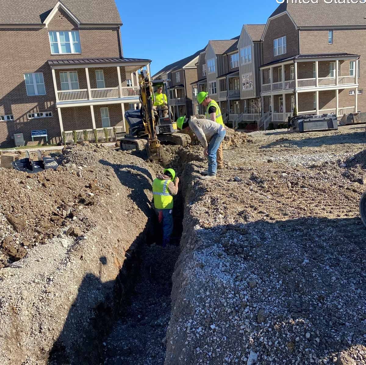 Construction workers excavating a trench near brick buildings; they wear safety vests and hard hats.