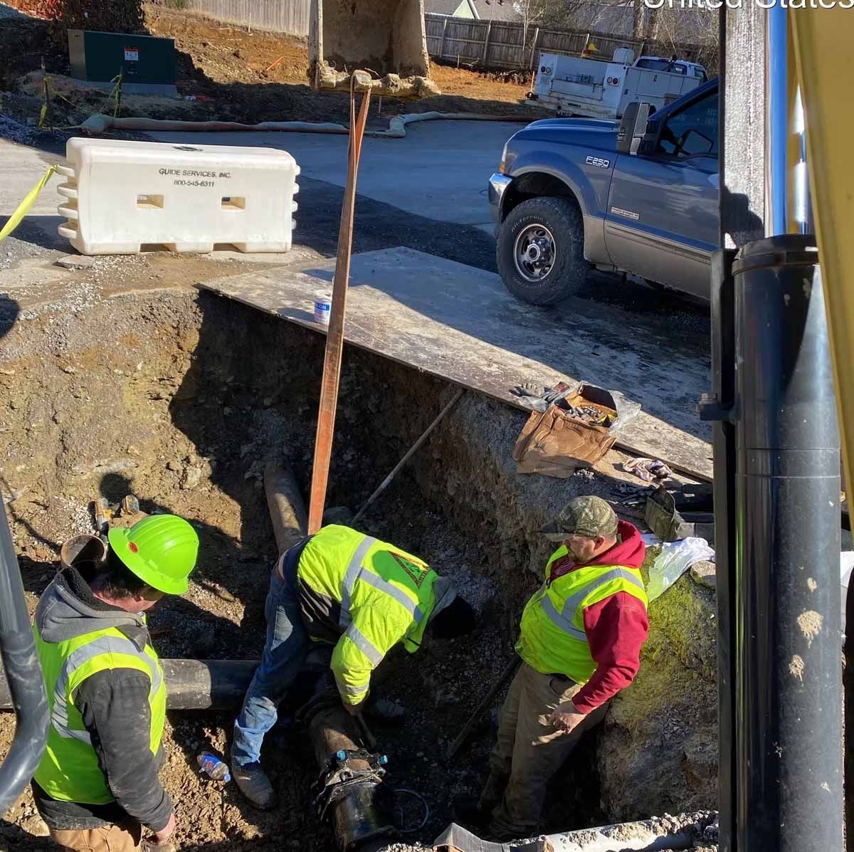 Three construction workers in vests and hard hats work on pipes in a roadside trench.