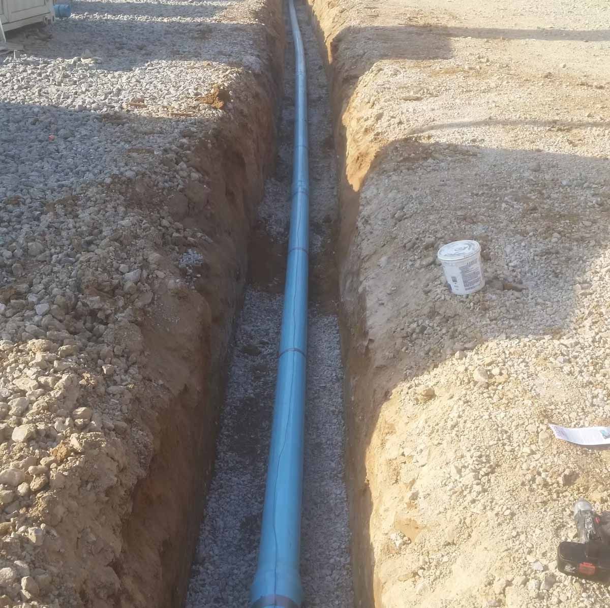 A long, blue pipe lies in a narrow trench filled with gravel, construction site setting.