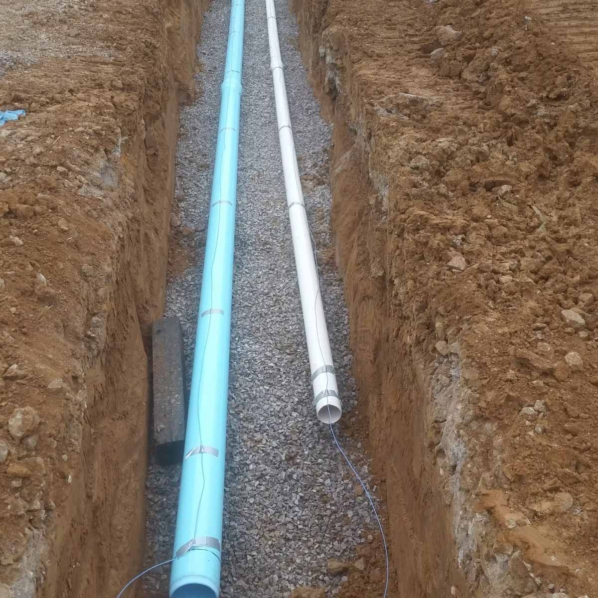 Trench with blue and white pipes, gravel base, in dirt.