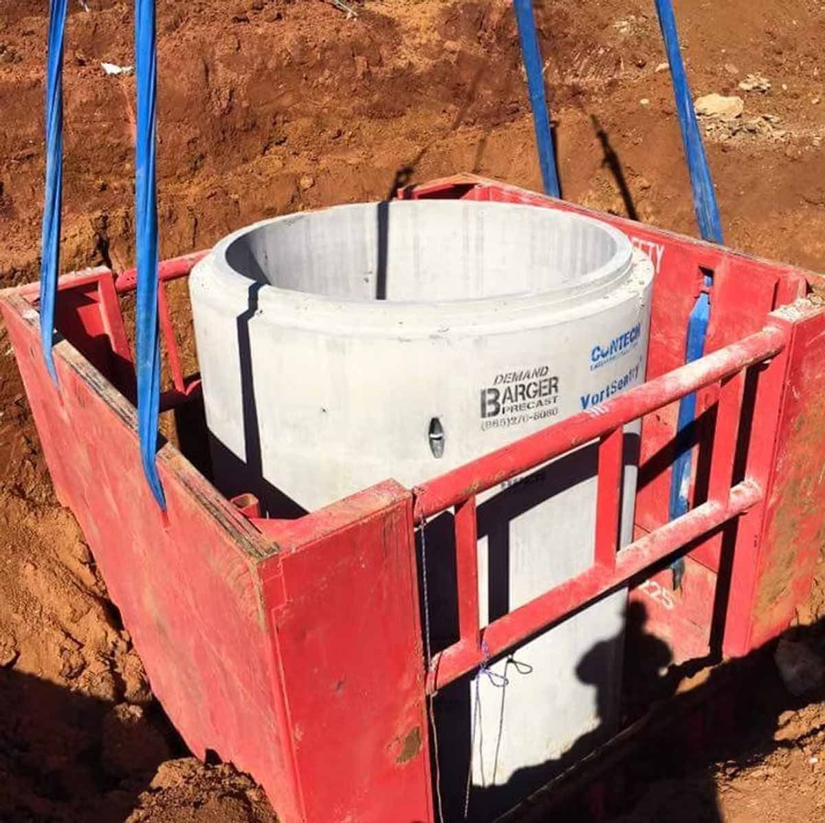 Concrete cylinder being lowered into an excavation pit using a red metal cage and blue straps.
