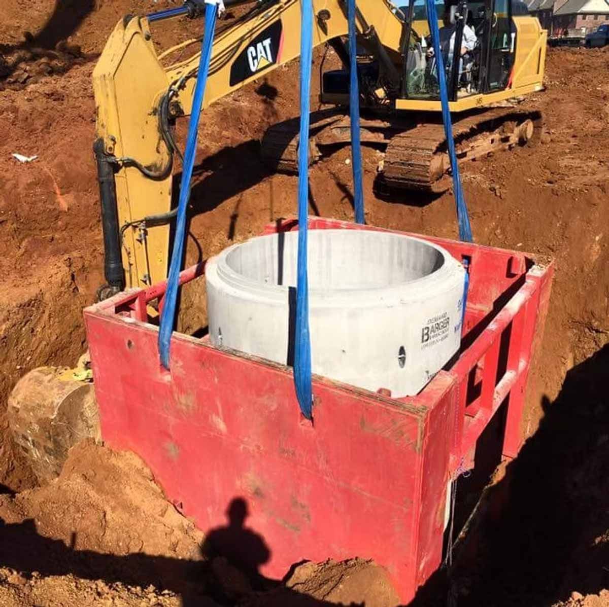 Yellow excavator lifting a concrete ring inside a red box in an earth pit.