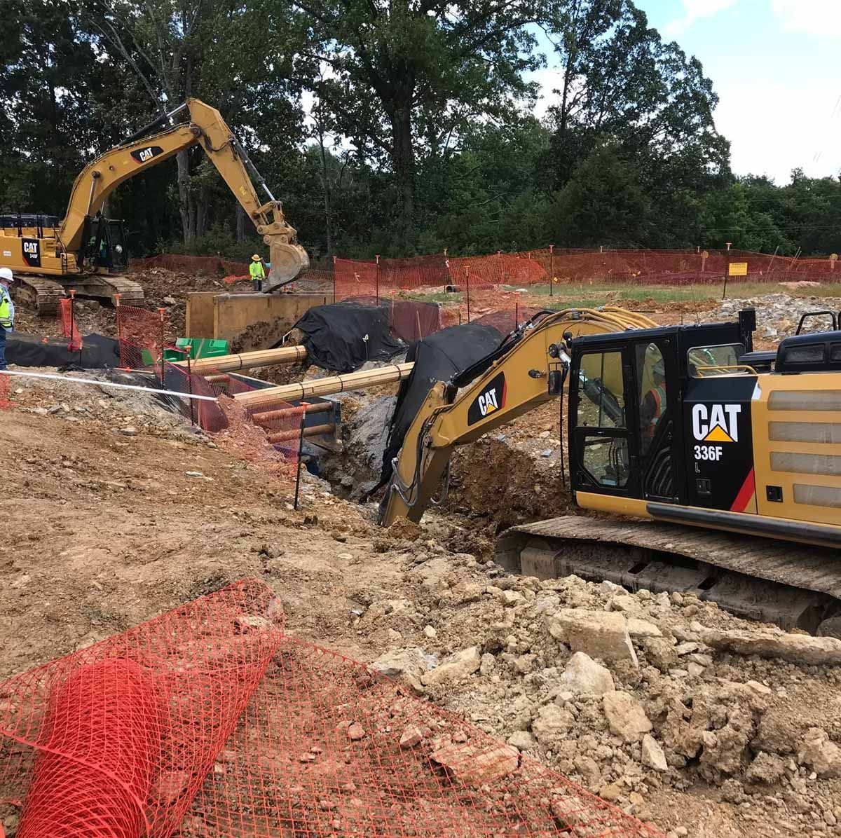 Two yellow CAT excavators digging in an earthwork construction site; red safety fencing.