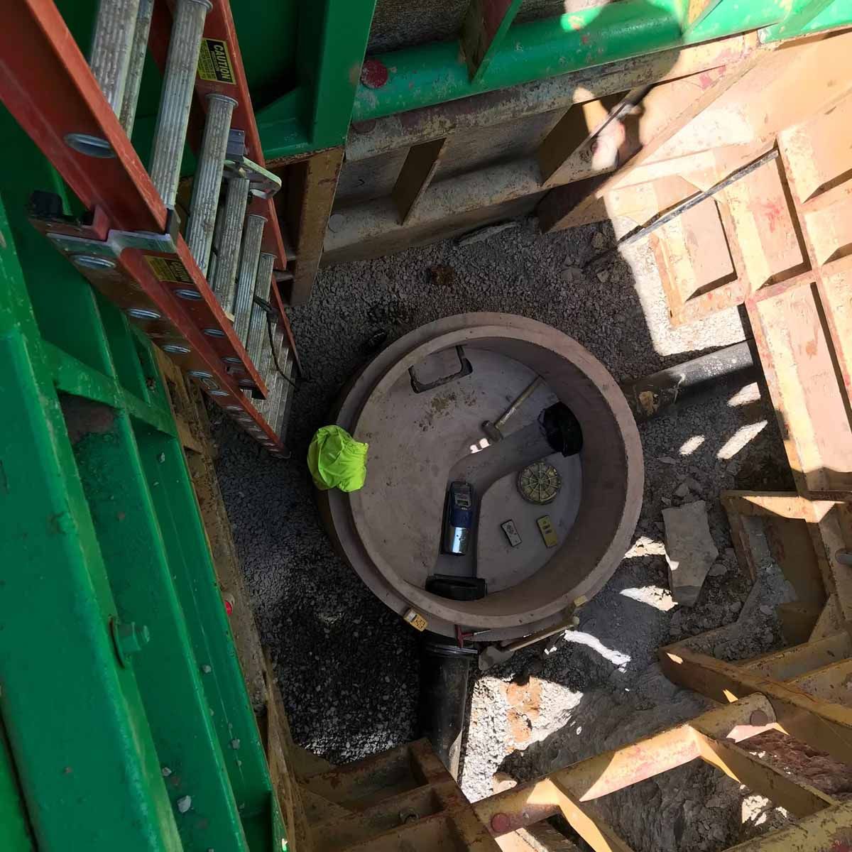 Open manhole in a construction site. A ladder, tools, and green fencing surround it.
