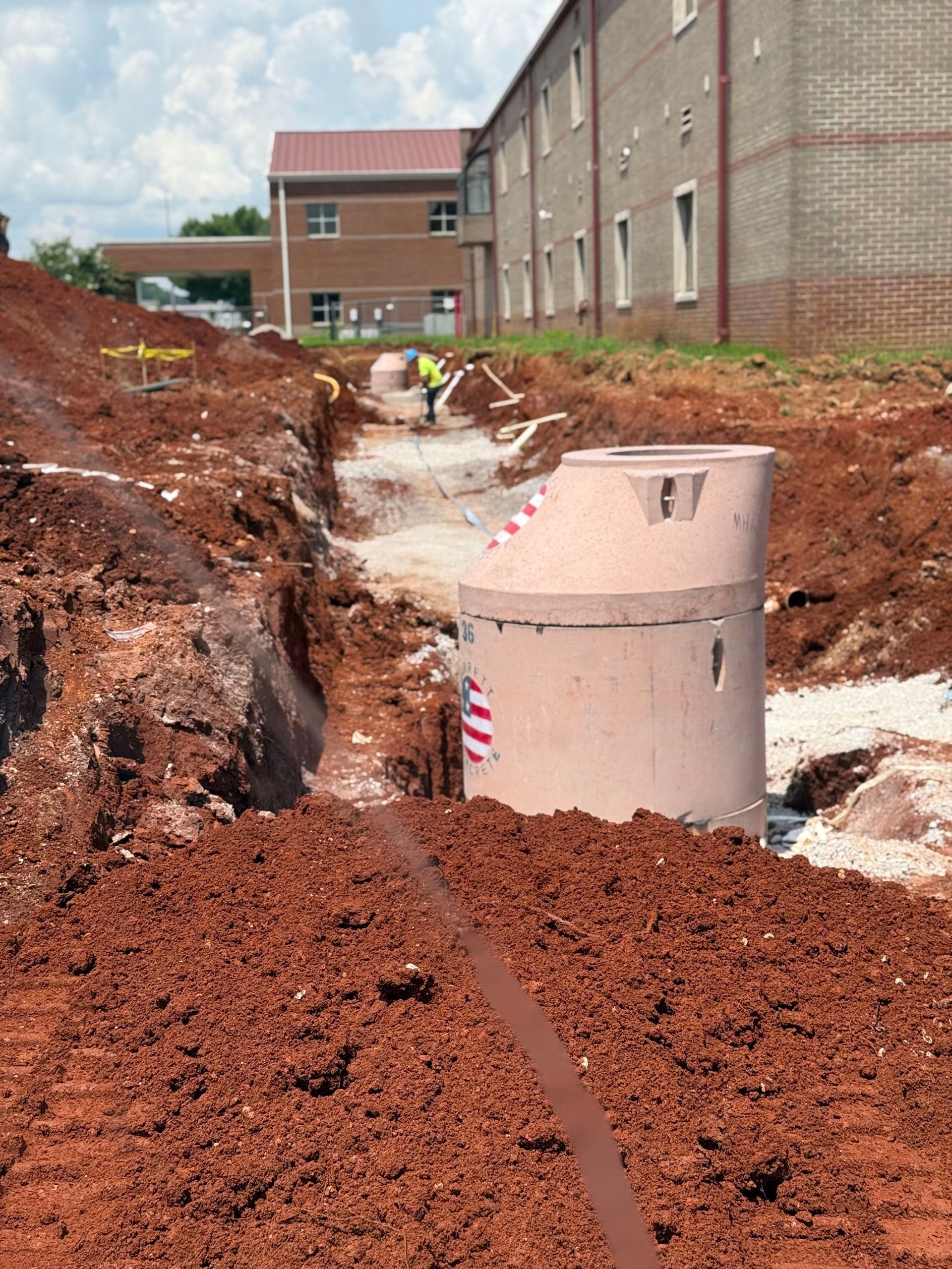 A construction site with a deep red-dirt trench and a concrete manhole structure near a brick building.