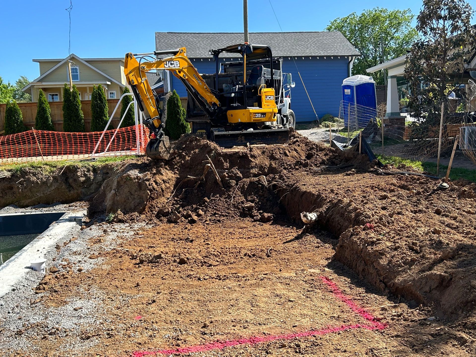 A yellow excavator parked behind a construction site with a freshly dug trench and pink markings on the ground.