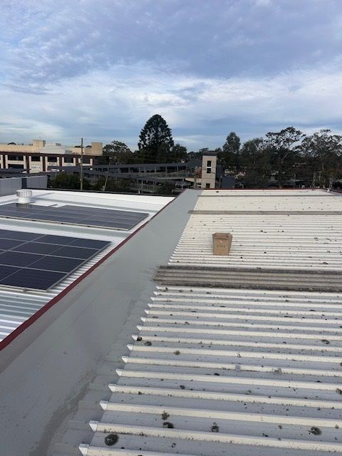 A rooftop with corrugated metal panels, solar panels, and a small box. Cloudy sky in the background with buildings and trees.
