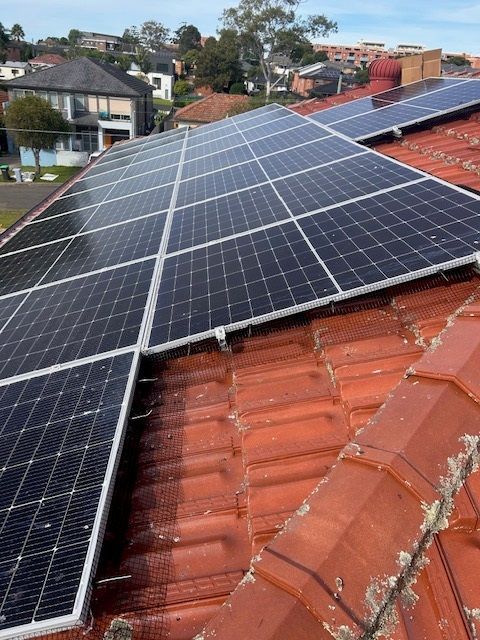 Solar panels installed on a red tile roof in a residential neighborhood. Blue sky visible.