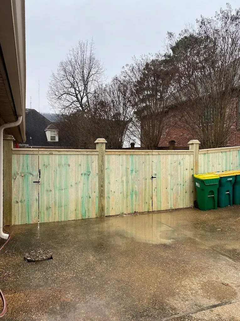 Wooden fence enclosing a wet concrete patio. Green and yellow trash bins sit on the right side.