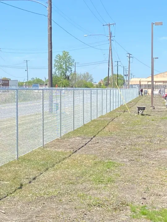 Chain link fence along a grassy area, with utility poles overhead and a building visible in the distance under a blue sky.
