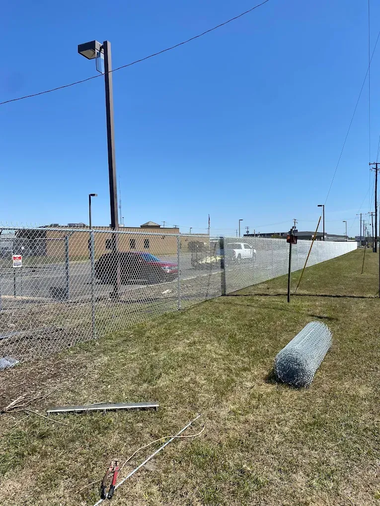 Chain-link fence with a building in the background. A roll of wire fencing sits on the grass. Clear blue sky.