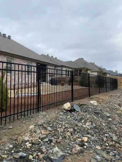 Black metal fence in front of multiple brick houses on a cloudy day, dirt and rocks in the foreground.