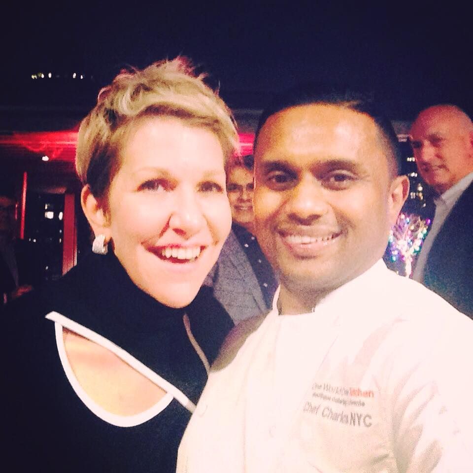 Woman and man smiling for a selfie; man in chef’s coat, other people in background.