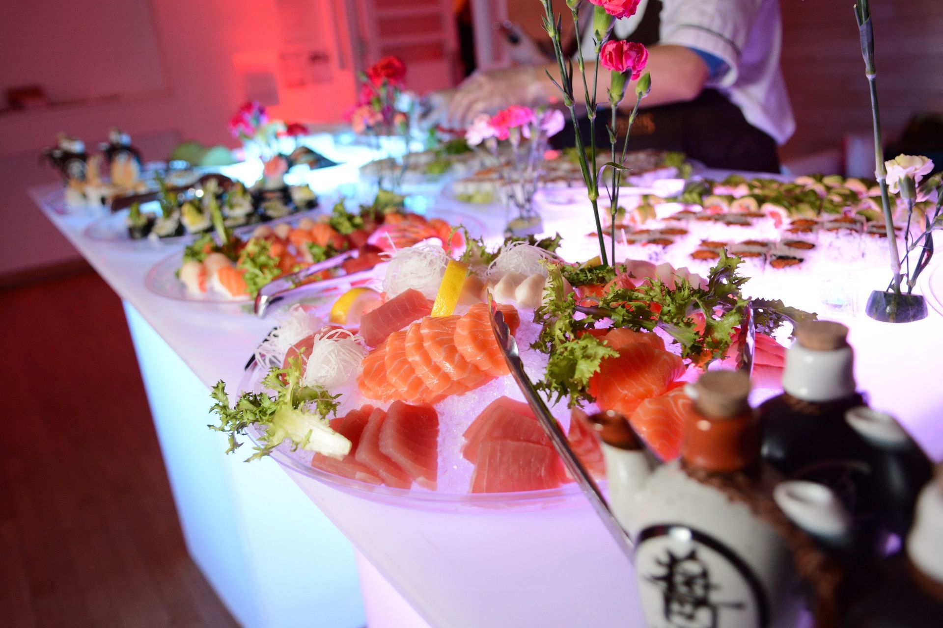 Sushi buffet with various dishes on ice, chef preparing food, and floral decorations.