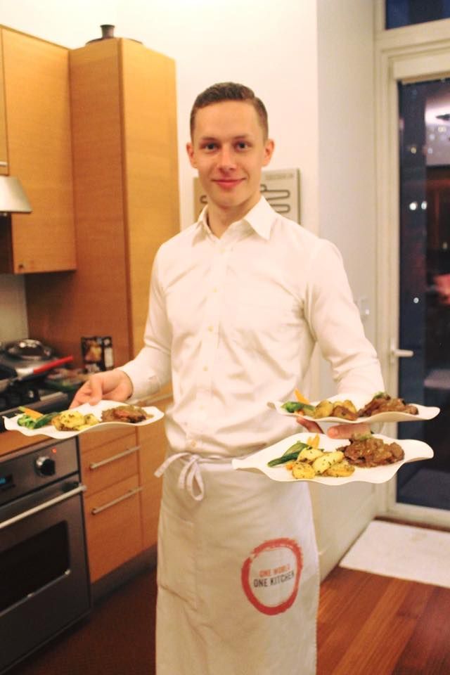 Man holding plates of food in a kitchen, wearing an apron, smiling.