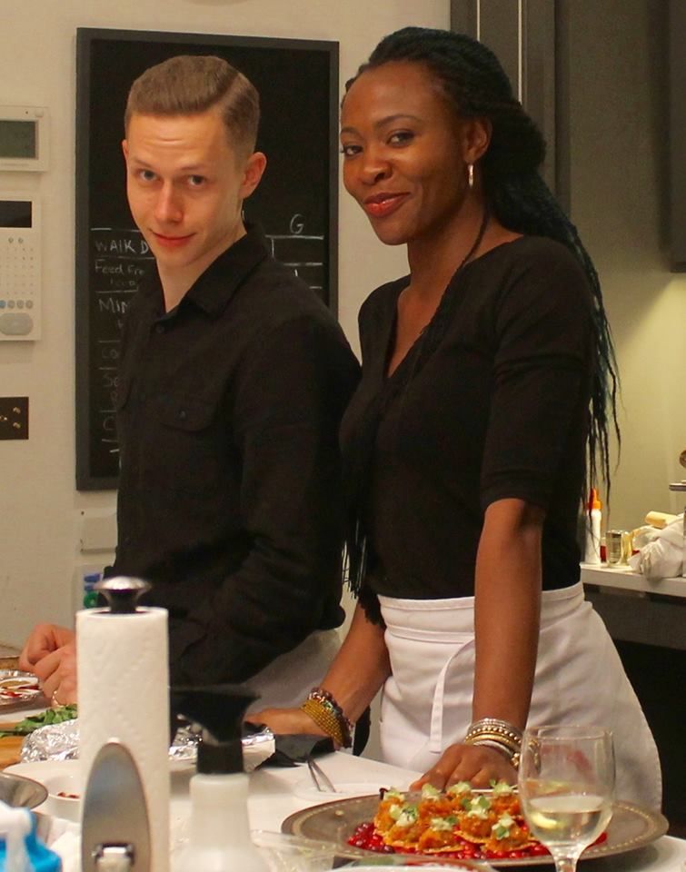 Two people in a kitchen smile, one with blue braids and a white apron, the other with a dark shirt.