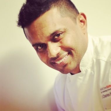 Chef smiling, wearing a white coat, leaning towards the camera.
