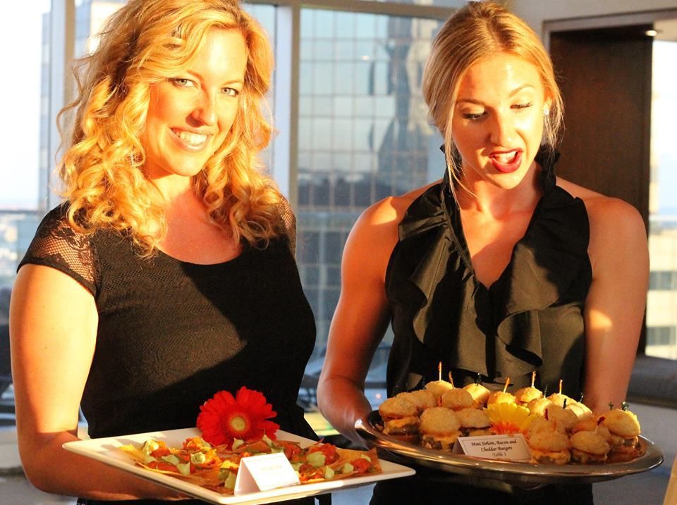 Two women holding trays of appetizers indoors with a city skyline in the background.