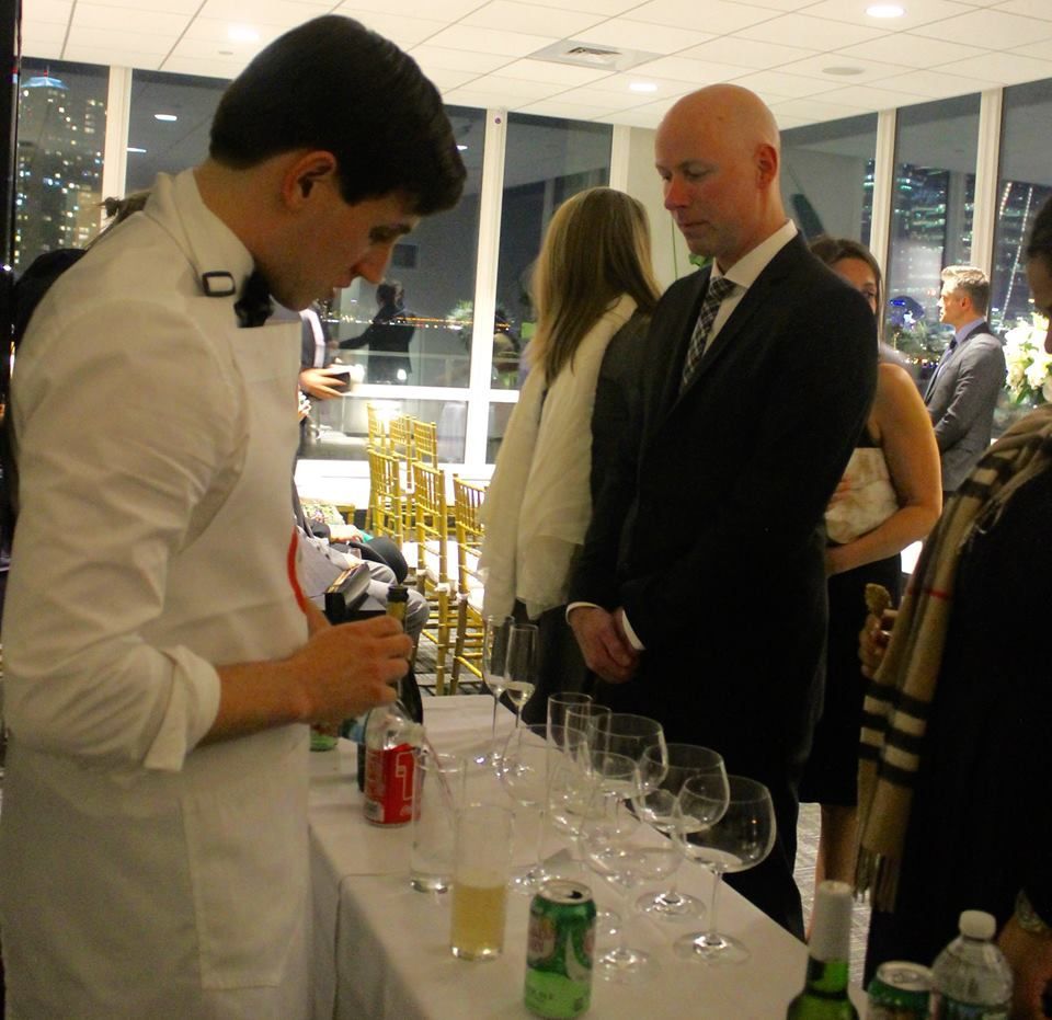 Bartender pours drinks at an event, guests watch. White apron, formal wear, glasses lined up.