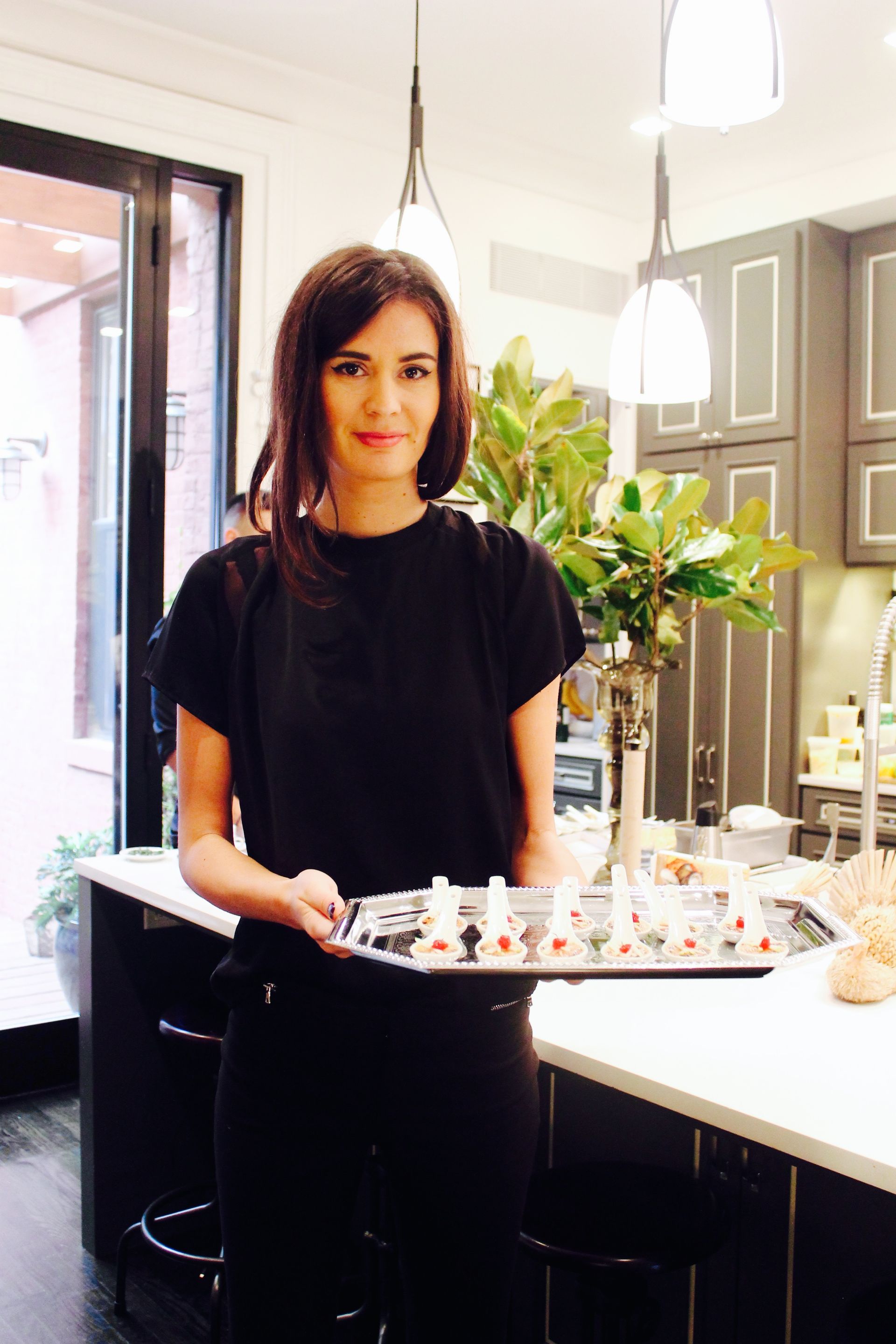 Woman holding a tray of food in a modern kitchen with dark cabinets and white countertops.