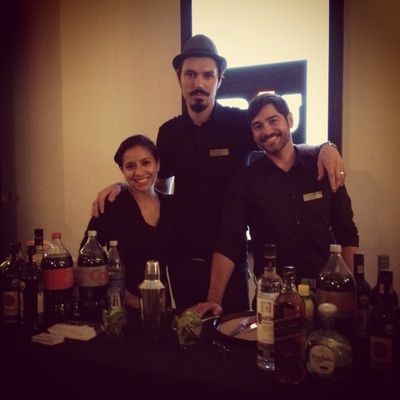 Three bartenders smiling behind a bar with bottles, one wearing a hat.