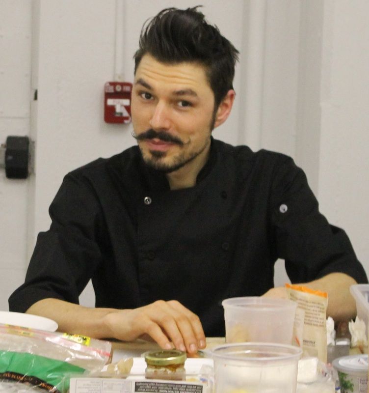 Man with dark hair and mustache wearing a black chef's coat, seated at a table with food items.