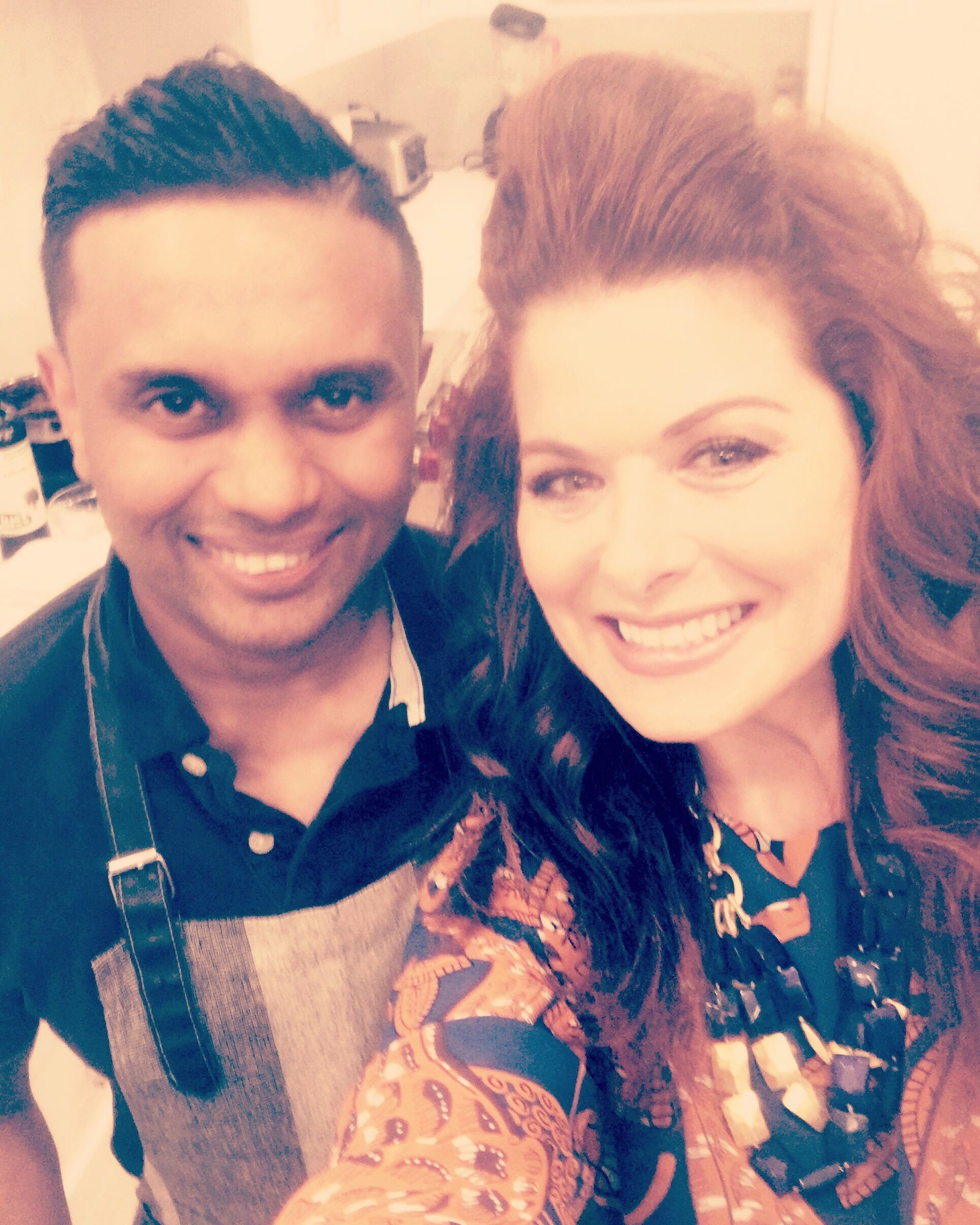 Two people smiling at the camera. A man wearing an apron, and a woman with red hair, in a kitchen setting.
