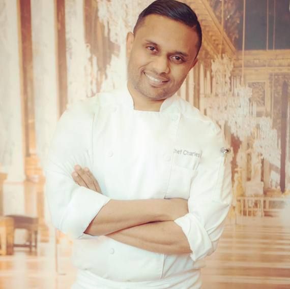Chef in white coat smiles, arms crossed, in front of ornate backdrop.