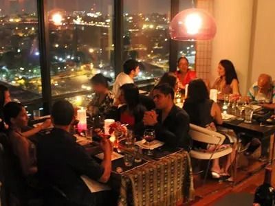 People dining at a table by a window with city lights. Lit by overhead light, wine glasses visible.