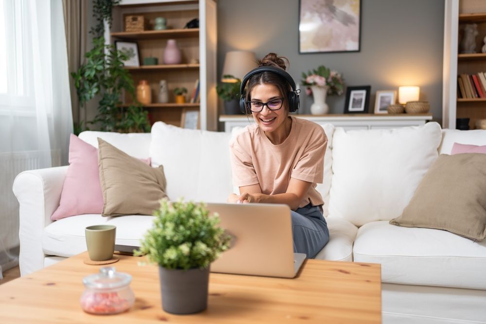 Woman wearing headphones, smiling at laptop on couch in a living room.