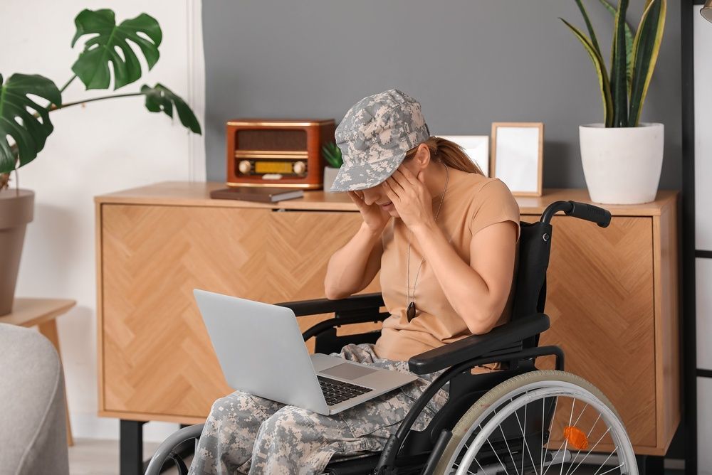 Woman in wheelchair, wearing camouflage, using laptop, resting hands on head, indoors.
