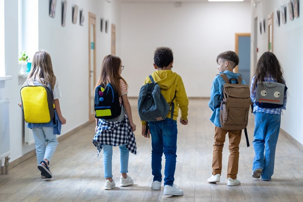 Five children walking down a school hallway, each wearing a backpack.