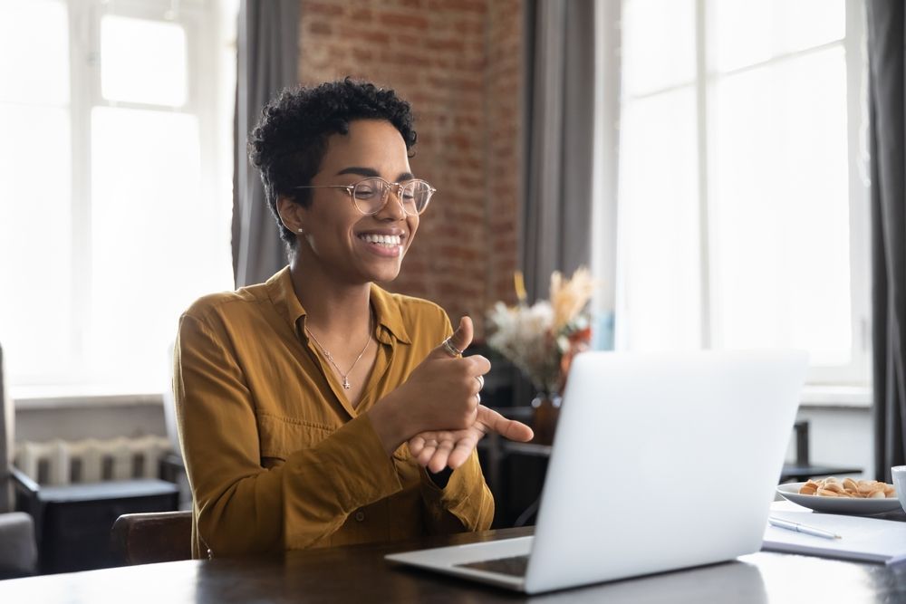 Woman using sign language on a video call. She smiles, sitting at a table with a laptop.