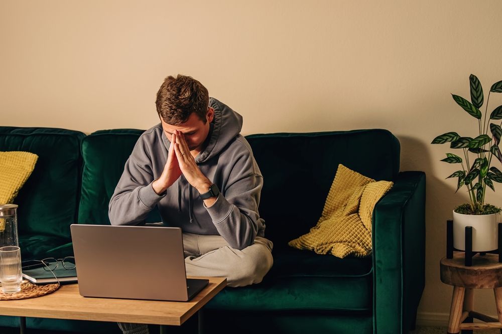 Man sits on couch with laptop, hands on face, looking stressed.