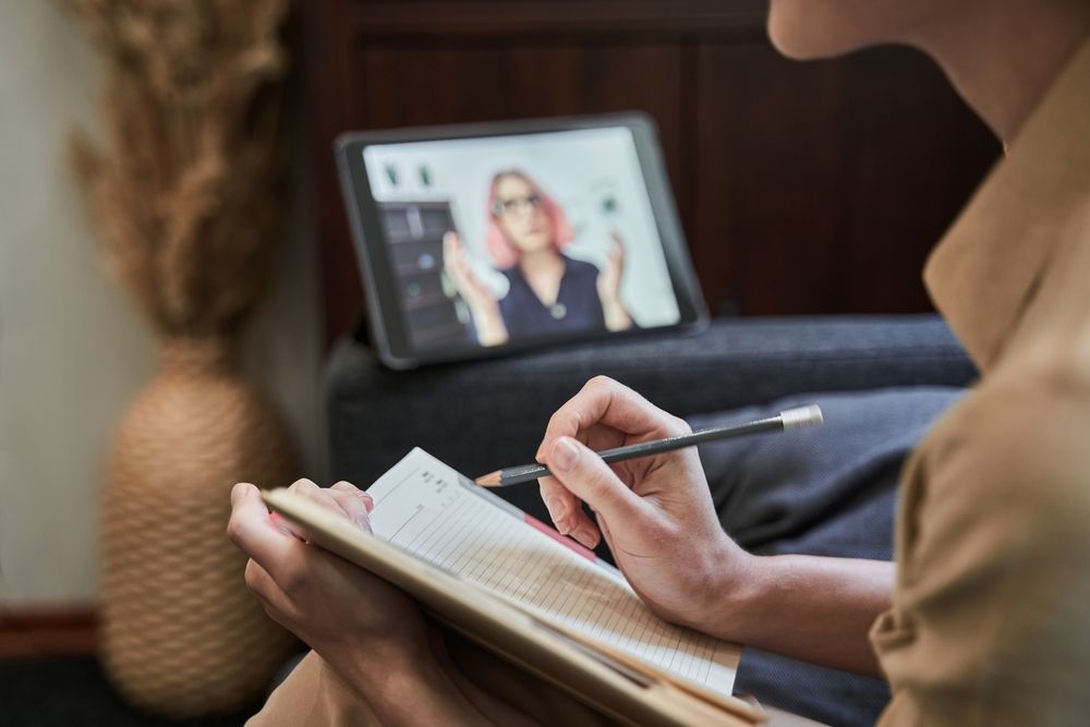 Person taking notes from a tablet showing a video call, in a home setting.