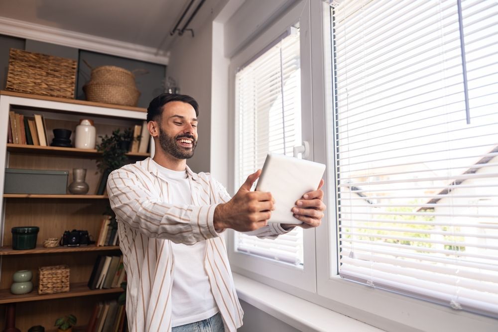 Man holding tablet near window, smiling, with a bookshelf in the background.