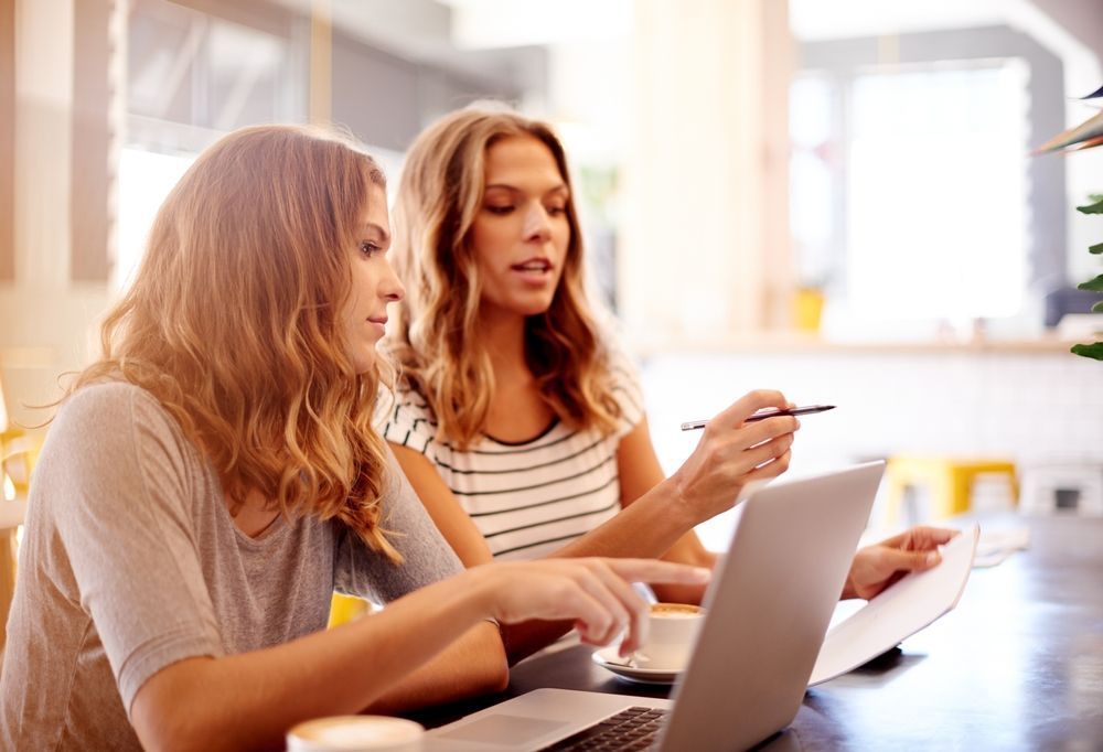 Two women at a table with a laptop, documents, and coffee, collaborating. One points, the other looks on.
